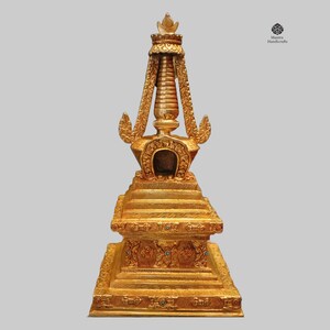 Könnte beinhalten: Eine goldfarbene Metallstupa mit filigranen Details und einem quadratischen Sockel. Die Stupa ist mit türkisfarbenen Edelsteinen verziert und hat eine Spitze mit einem Abschluss.
