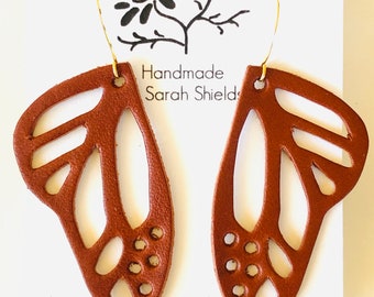 Puede incluir: Pendientes de mariposa de cuero marrón con recortes y ganchos dorados. Los pendientes están hechos a mano por Sarah Shields y están disponibles para comprar en su tienda Etsy.