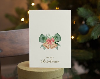 Puede incluir: Una tarjeta de felicitación rectangular de color blanco roto con una ilustración en acuarela de dos campanas doradas atadas con una cinta roja y un gran lazo verde, acentuadas con agujas de pino. El texto MERRY christmas está impreso en oro.