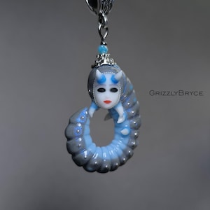 May include: A unique pendant with a fantasy-inspired design. The pendant depicts a figure with a white mask, blue horns, and a gray and blue body. It hangs from a silver-toned bail and a white cord. The text "GrizzlyBryce" is visible. The pendant is a piece of jewellery.