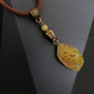 May include: A gold pendant necklace with a brown leather cord. The pendant features a clear, yellow, and brown stone with small, red flecks. The stone is attached to a gold chain with a small, gold flower charm.