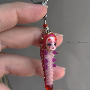 Op de afbeelding: Een hanger aan een ketting met een gebeeldhouwd wezen met een bleek gezicht, rode hoorns en donkere markeringen rond de ogen. Het lichaam is roze en gesegmenteerd met paarse uitsteeksels, in een rood-gouden schaal. Tekst: GRIZZLYBRYCE.
