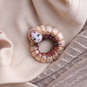 May include: A distinctive, circular brooch with a stylized face and red accents, set on a cream-coloured body. The brooch is ring-shaped, with brown and gold detailing. The background is a beige fabric.