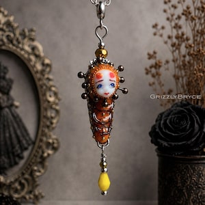 Peut inclure: Un collier pendentif artisanal avec un charmant pendentif en forme de poupée. Le pendentif présente un visage détaillé avec des yeux bleus et des accents rouges, sur un corps brun segmenté. Le collier comprend une chaîne argentée et une perle jaune en forme de goutte.