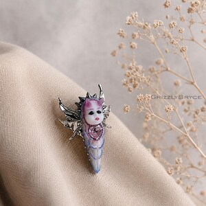 May include: A silver-toned brooch featuring a central pale face with purple horns, dark eyes, and red lips. The figure is framed by metallic wings and spikes, with a heart-shaped purple gemstone set below the face. The text GRIZZLYBRYCE is visible.