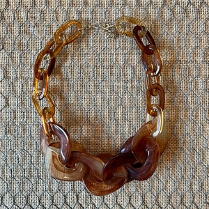 Puede incluir: Un collar de resina marrón y ámbar con eslabones grandes e interconectados. El collar está hecho de resina gruesa y opaca y tiene un acabado pulido.