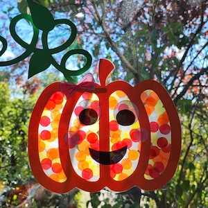 Peut inclure: Une décoration de fenêtre en forme de citrouille avec un contour orange vif, une tige verte avec des vrilles, et un centre translucide rempli de confettis circulaires rouges, orange et jaunes. Elle a un visage souriant et un petit nœud rose.