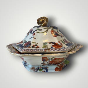 Puede incluir: Un plato de servir antiguo con tapa, adornado con un motivo floral en naranja, azul y verde sobre fondo blanco. La tapa está rematada con un pomo dorado. El plato presenta un borde festoneado, típico de la vajilla vintage.