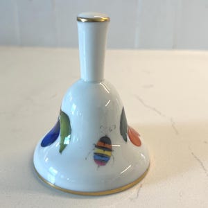 May include: White porcelain bell with a gold rim and handle. The bell features a colorful insect illustration and floral accents in blue, green, and orange. A decorative piece for home decor.