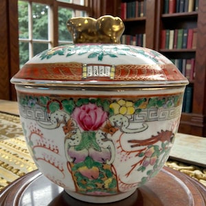 May include: A decorative porcelain bowl with a lid, featuring a vibrant floral design in pink, yellow, and green. The rim is accented with red and gold, and the lid has a gold-colored handle. The bowl is set on a wooden stand.