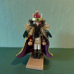 May include: A wooden king nutcracker figurine. The king wears a gold-trimmed green and purple cape, a gold crown with a red top, and black boots. The figurine stands on a small wooden base. The king has a black beard and mustache.