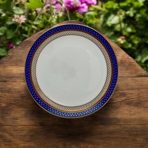 May include: A white ceramic plate with a blue and gold rim. The plate has a decorative pattern of dots and lines. The plate is sitting on a wooden surface.