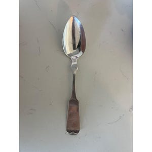 May include: A silver spoon with an oval bowl and a long, slender handle. The handle has a decorative design near the bowl and a rectangular end with a curved bottom. The spoon is reflective, showing the surrounding environment.