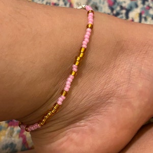 May include: A pink and gold beaded ankle bracelet. The bracelet is made of small, round beads that are strung together on a thin cord.