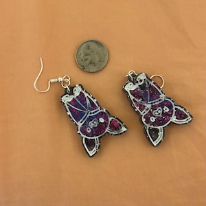 Op de afbeelding: Een paar oorhangers met een vleermuisontwerp van paarse en zwarte glitter met witte accenten. De oorbellen hebben zilveren haakjes.