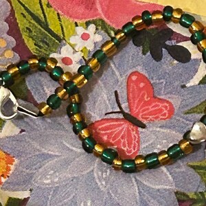 Könnte beinhalten: Ein grünes und goldenes Perlenarmband mit einem silbernen Kleeblatt-Anhänger. Das Armband hat einen silbernen Verschluss.