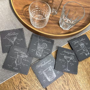 Può includere: Un set di sei sottobicchieri quadrati in ardesia con illustrazioni bianche di cocktail classici, tra cui Martini e Margarita. Sono visibili anche bicchieri trasparenti e un vassoio di legno.