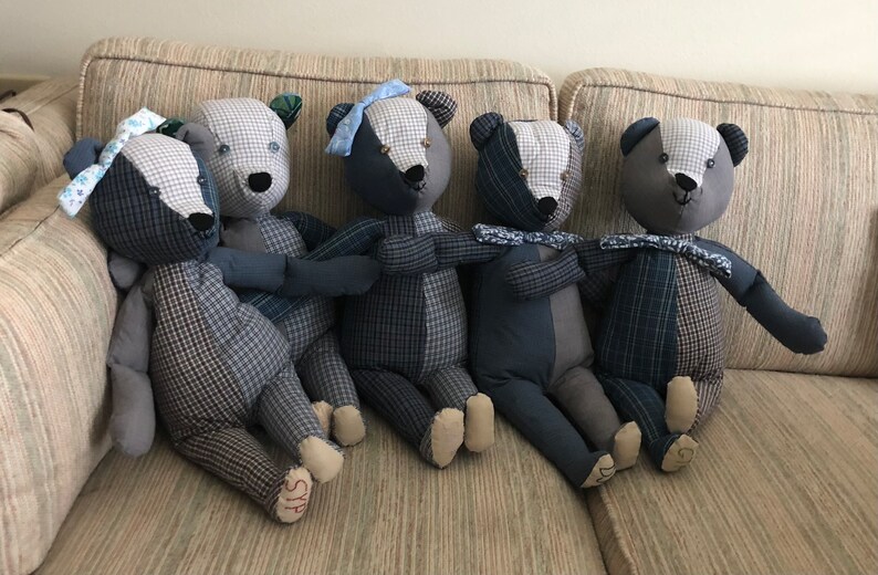 May include: Five patchwork teddy bears in blue and grey plaid fabric sitting on a beige sofa. The bears have black eyes and noses and are all wearing blue and white ribbon bows.