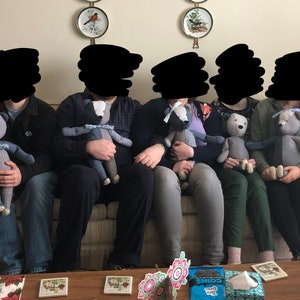 May include: Five people are sitting on a sofa, each holding a grey and blue patchwork teddy bear. The bears have different facial expressions and are all wearing a blue bow tie.