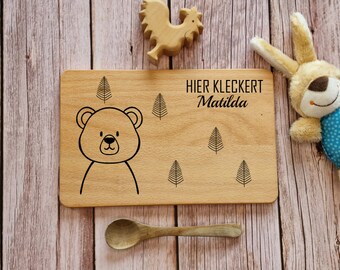 May include: A wooden cutting board with a bear illustration and the text "HIER KLECKERT Matilda". The board is surrounded by a wooden surface and a wooden toy rooster and a stuffed bunny.