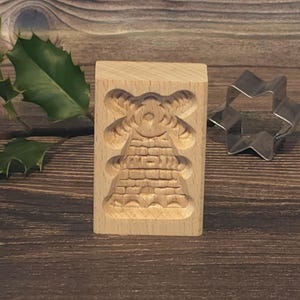 May include: A wooden cookie cutter shaped like a Christmas tree with a detailed design. The cutter is on a wooden surface.