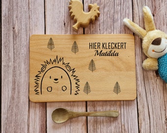 May include: A wooden cutting board with a hedgehog design and the text "HIER KLECKERT Matilda". The board is surrounded by small trees and is on a wooden surface.
