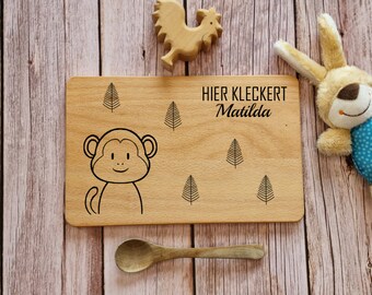 May include: A wooden cutting board with a monkey illustration and the text "HIER KLECKERT Matilda". The board also features a few tree illustrations. A wooden spoon and a stuffed bunny are also in the image.