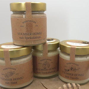 May include: Four glass jars with gold lids and brown paper labels. The labels have the text "Wabenwerk" and "Volmser Honig" with different flavor variations: "Spekulatius", "Zimt", "Tonkabohne", and "Vanille".