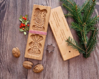 Könnte beinhalten: Zwei Holz-Ausstechformen, eine in Form eines Lebkuchenhauses und die andere in Form eines Weihnachtsbaums, mit einem rot-weiß karierten Band um die Lebkuchenhaus-Ausstechform gebunden. Die Ausstechformen befinden sich auf einer Holzoberfläche mit einem Zweig Stechpalme und einer sternförmigen Ausstechform.
