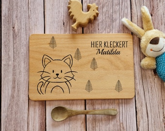 May include: A wooden cutting board with a cat illustration and the text "HIER KLECKERT Matilda."  The board is surrounded by wooden toys, including a rooster and a bunny.