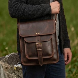 May include: A dark brown leather messenger bag with a fold-over flap, a front pocket, and a vertical strap secured by a brass-toned metal buckle. The bag features visible stitching along the edges and two small brass rivets on the top flap.