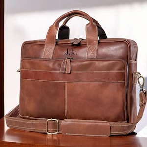 Op de afbeelding: Een bruine leren aktetas met een handvat aan de bovenkant en een afneembare schouderriem. De aktetas heeft een voorvak, een ritssluiting en de initialen "J.K." zijn op de bovenkant geëmbosseerd. De tas staat op een houten oppervlak.