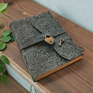 Könnte beinhalten: Ein graues Lederjournal mit geprägtem Blumenmuster. Das Journal hat einen Klappenverschluss, der mit einem herzförmigen Schloss und einem kleinen Schlüssel gesichert ist. Die Seitenränder sind goldfarben.