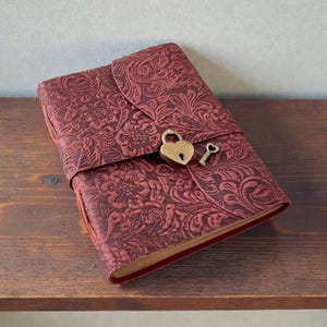 May include: A burgundy journal with a floral embossed design. The journal has a heart-shaped lock and key closure. The journal is resting on a wooden surface.