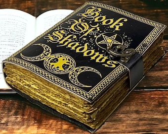 May include: A black leather-bound journal with gold accents and the words "Book of Shadows" on the cover. The journal has a pentagram and a crescent moon design.
