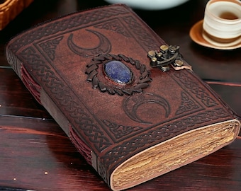 May include: A brown leather journal with a Celtic knot design and a triple moon symbol. The journal has a blue gemstone and a brass clasp.