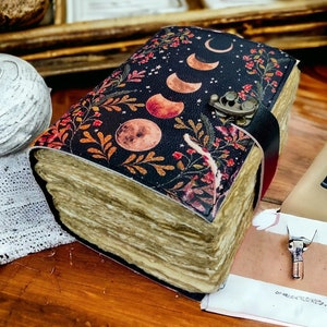 Puede incluir: Un diario encuadernado en cuero negro con un dise&ntilde;o floral y lunar. La portada presenta un patr&oacute;n repetitivo de hojas, flores y lunas crecientes. El diario est&aacute; cerrado y tiene un cierre de metal.