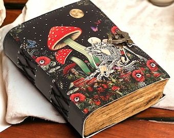 Peut inclure: Un journal en cuir noir avec une illustration colorée de deux squelettes qui s'embrassent dans un décor de forêt. Le journal est doté d'une serrure et d'une clé et présente des champignons, des fleurs et des papillons.