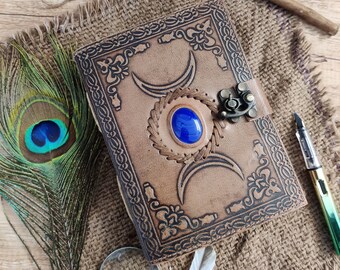 May include: A brown leather journal with an embossed design, a blue gemstone, and a metal clasp. The journal is surrounded by a peacock feather, a magnifying glass, a feather, a pencil, and a pen.