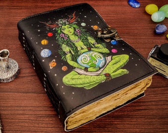 May include: A black leather journal with a clasp, featuring a green goddess illustration holding a planet. The journal is surrounded by a quill pen, inkwell, and decorative stones. The pages have a vintage, aged appearance.