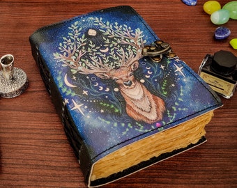 Puede incluir: Un diario encuadernado en cuero azul con una ilustración detallada de un ciervo con cuernos rodeado de estrellas, lunas y hojas. El diario tiene un cierre de metal y está cerrado.