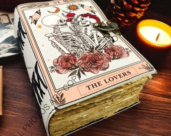 Op de afbeelding: Een wit lederen gebonden dagboek met een roze en witte illustratie van twee skeletten die elkaars hand vasthouden. De illustratie is omringd door bloemen en de tekst "THE LOVERS" is onder de afbeelding gedrukt.