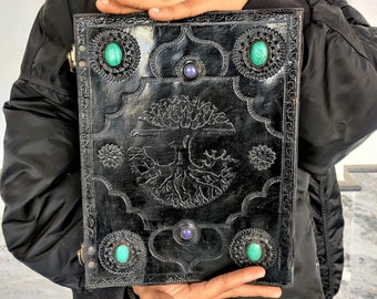 May include: A black leather journal with a tree of life design and turquoise and purple gemstones. The journal has a flap closure and a metal clasp.