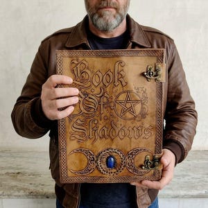 May include: A light brown leather-bound book with the title "Book of Shadows" embossed on the cover. The cover features a pentagram, crescent moons, and a blue gemstone. The book has metal clasps.