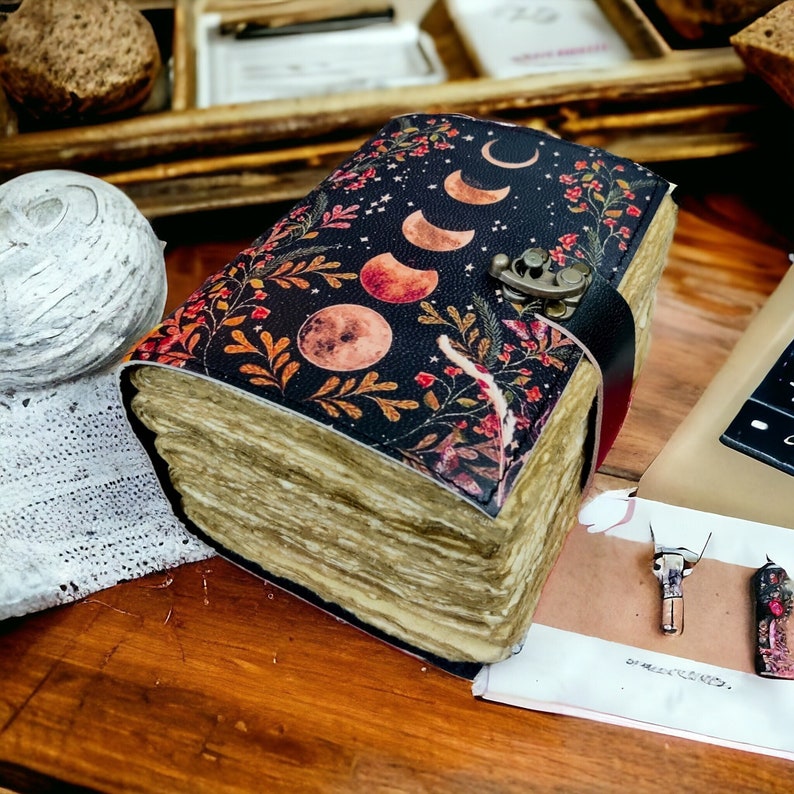 Puede incluir: Un diario encuadernado en cuero negro con un dise&ntilde;o floral y lunar. La portada presenta un patr&oacute;n repetitivo de hojas, flores y lunas crecientes. El diario est&aacute; cerrado y tiene un cierre de metal.