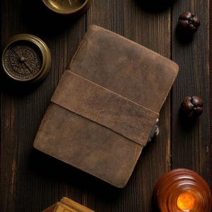 May include: A brown leather journal with a strap closure sits on a dark wooden surface, surrounded by antique brass compasses and decorative items. The journal's cover has a textured appearance. The scene evokes a vintage, travel-themed aesthetic.