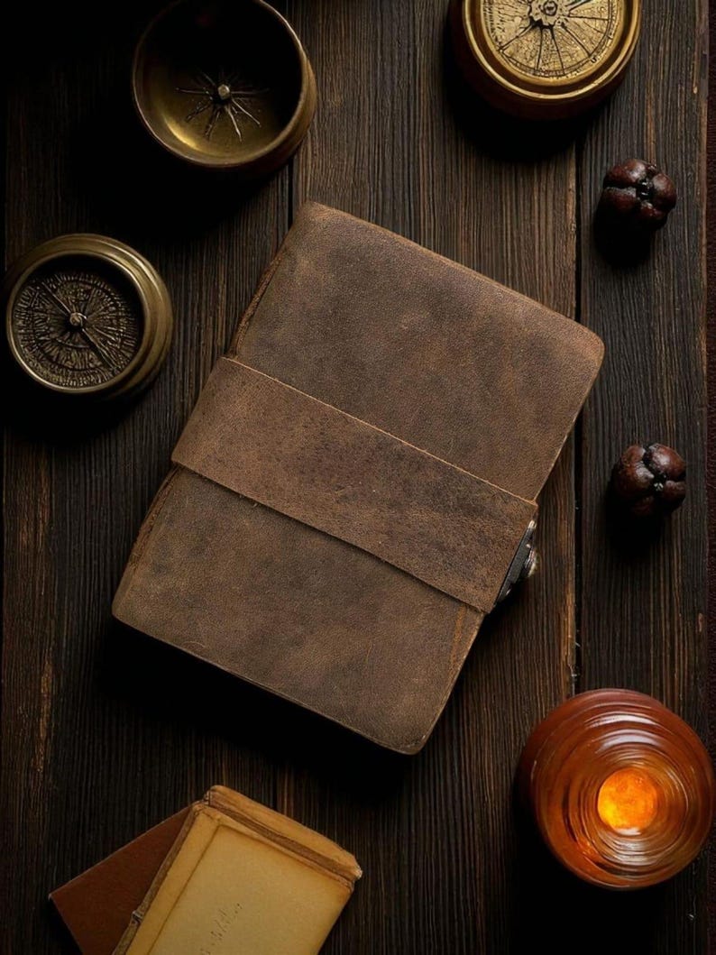 May include: A brown leather journal with a strap closure sits on a dark wooden surface, surrounded by antique brass compasses and decorative items. The journal's cover has a textured appearance. The scene evokes a vintage, travel-themed aesthetic.