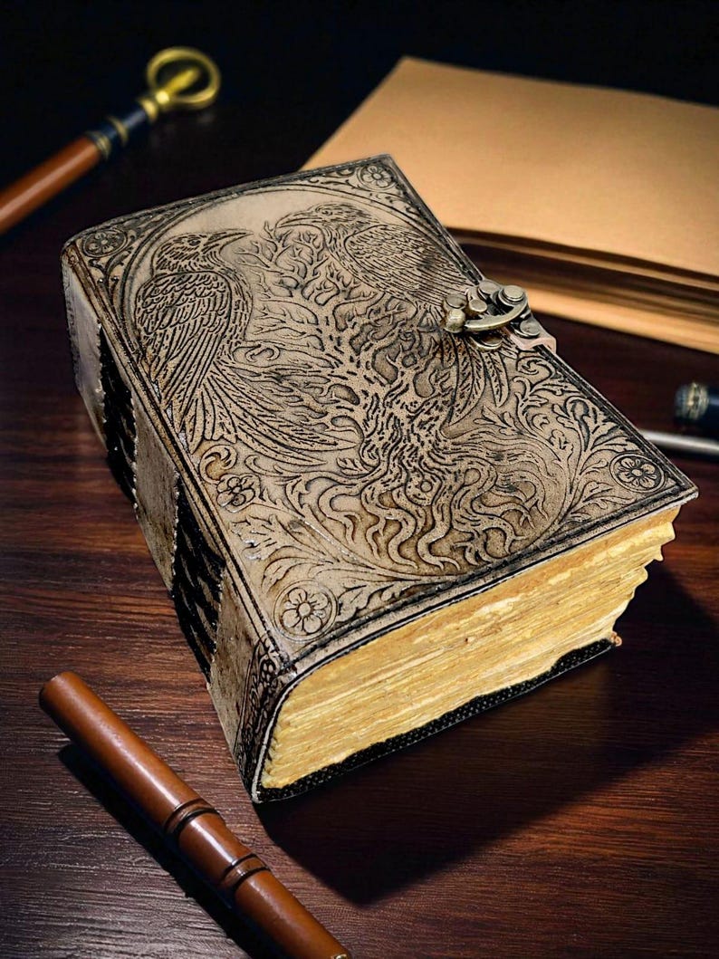 Puede incluir: Un diario de cuero de estilo vintage con un dise&ntilde;o en relieve que presenta dos aves y un &aacute;rbol. El diario tiene un cierre de metal y p&aacute;ginas envejecidas y amarillentas. Una pluma de madera descansa cerca, sugiriendo un tema de escritura.