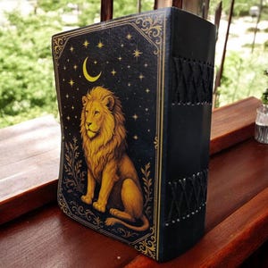 May include: A black journal with a golden lion illustration on the front cover. The cover also features a crescent moon, stars, and decorative gold trim. The spine is black with a stitched design.