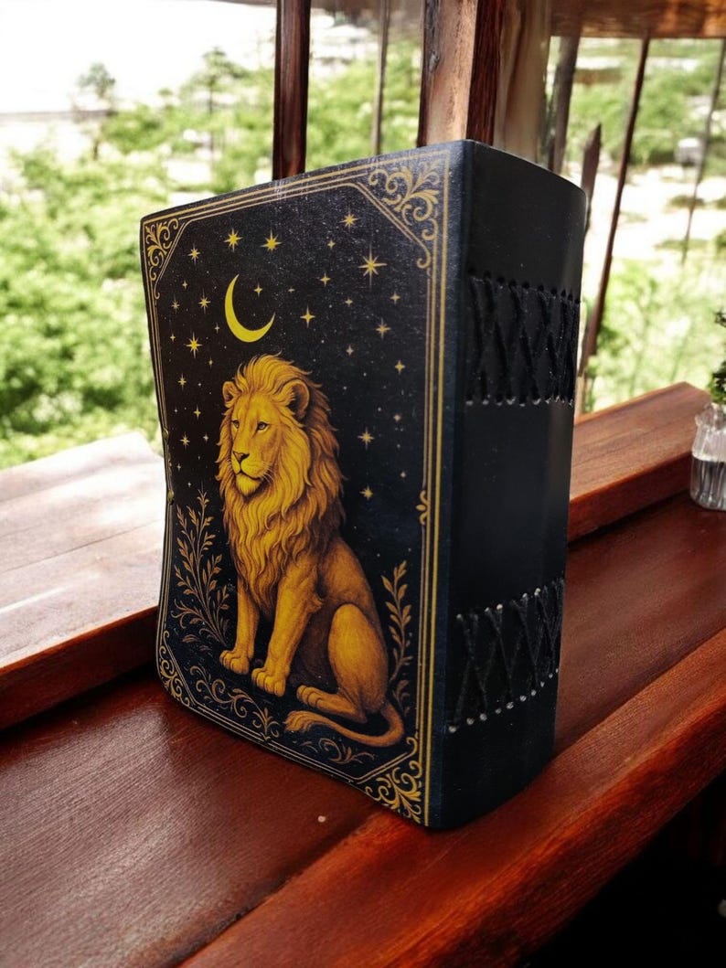 May include: A black journal with a golden lion illustration on the front cover. The cover also features a crescent moon, stars, and decorative gold trim. The spine is black with a stitched design.